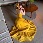 A woman wearing a bright yellow sleeveless maxi dress with a ruffled hem, kneeling on a gray carpeted floor in a modern living room setting.
