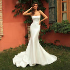 Woman in a white strapless gown standing outdoors against a red wall with greenery.