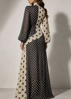 A woman modeling a long black and beige polka dot dress from the back, featuring sheer long sleeves and a high neckline, set against a neutral studio background with a soft shadow.