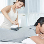 Woman using a handheld device on a man lying on a bed in a bright room.