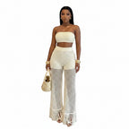 A woman stands in a white arched alcove wearing a cream-colored crochet two-piece set consisting of a strapless crop top and high-waisted, wide-leg fringe pants. She is accessorized with gold bracelets, a woven straw handbag, and gold sandals.