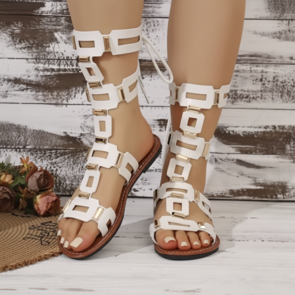 White strappy flat sandals with geometric square cutouts and lace-up ankle ties, shown on a model's feet against a rustic whitewashed wood plank background.