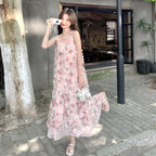 A woman stands outdoors on a paved street in front of a building, wearing a long light pink sleeveless dress with floral embroidery and 3D flower details. She is holding a small white quilted handbag and wearing white sandals.