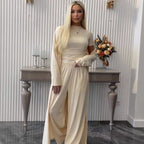 A woman with long blonde hair models a matching beige long-sleeve top and wide-leg trousers set, standing in front of a white ribbed wall with a decorative console table and dried flowers.