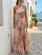 A woman models a one-shoulder maxi dress with a pink base and green floral print, standing on a cobblestone street in a Mediterranean village setting.
