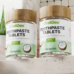 Two jars of OralGos toothpaste tablets on a bathroom shelf with a plant and mirror in the background.