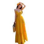 A woman wearing a bright yellow maxi dress with a deep V-neckline and a gathered waist stands beside a swimming pool. She is holding a woven straw bag and has a yellow flower in her hair, set against a background of wooden shutters and a tiled pool.