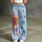 Light wash blue denim jeans with colorful hand-painted style rose designs on the legs, shown on a model from the waist down against a gray concrete floor.