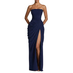 A woman models a strapless navy blue floor-length gown with a ruched bodice, draped skirt detail, and a high front slit, photographed against a clean light gray studio background.