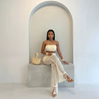 A woman sits on a concrete bench in a minimalist white room with an arched alcove, wearing a cream-colored two-piece knit set consisting of a strapless tube top and matching fringed maxi skirt. She is accessorized with a woven straw handbag and strappy sandals.