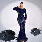 Woman in a long, dark blue dress with intricate patterns standing in front of rocks on a reflective surface.