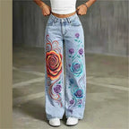 A person wearing light-wash blue jeans with large colorful floral prints, standing against a gray concrete wall.
