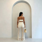 A woman stands with her back to the camera wearing a cream-colored crochet two-piece set consisting of a strapless bandeau top and high-waisted fringed wide-leg pants. She is holding a small woven straw bag in front of a minimalist white wall with an arched alcove.