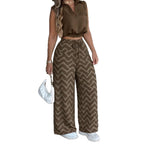 A woman modeling a two-piece clothing set featuring a brown satin crop top and matching wide-leg pants with a brown and beige wavy chevron pattern, styled with white sneakers and a white handbag against a plain white background.