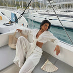A woman posing on a white yacht in a marina, wearing a matching white textured two-piece set consisting of a long-sleeve cropped tie-front top and wide-leg pants. She is holding a woven straw tote bag, with a matching folded fan on the deck and boats in the background.