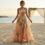 A woman stands on a sandy beach wearing a one-shoulder formal gown with a floral print in soft pink and green tones. The dress has a high slit, a fitted bodice, and a full flowing skirt.