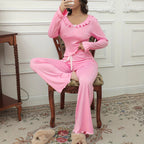 A woman wearing a pink two-piece pajama set with long sleeves and flared pants, sitting on a wooden chair in a decorated room.