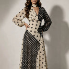 A woman models a long-sleeve midi dress with a split black and beige polka dot pattern, a wrap-style V-neckline, and a high neck, set against a neutral studio background with soft shadows.