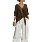 A woman wearing a brown short-sleeve V-neck top with a twisted knot detail at the waist, paired with wide-leg white pants, layered necklaces, and holding a woven clutch, set against a plain white background.