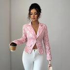 A woman wearing a pink floral-patterned blazer and white pants, holding a coffee cup and a small handbag against a plain light gray wall.