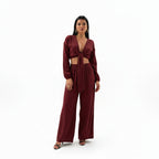 A woman modeling a matching burgundy ribbed two-piece set consisting of a long-sleeve wrap-style crop top and wide-leg pants, posed dynamically against a solid white background.