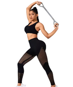 A woman in a black sports bra and black leggings with fishnet side panels poses while holding a gray resistance band against a white studio background.