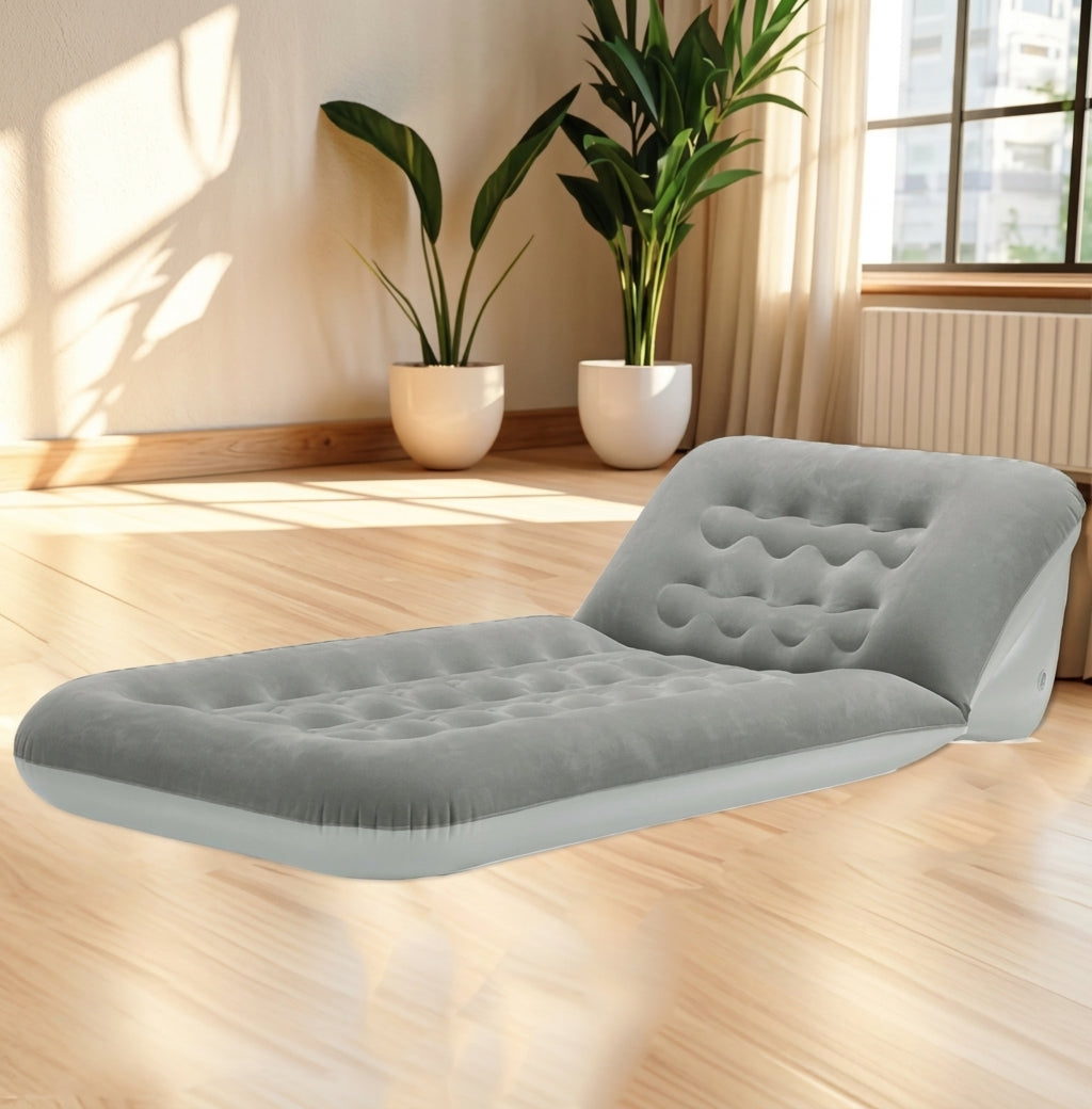 A light gray inflatable air mattress bed with a tufted headrest, shown on a light wood floor in a bright room with potted plants and a window.