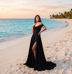 A woman stands on a sandy beach wearing a floor-length black off-the-shoulder gown with a high thigh slit. The background features turquoise ocean water, a soft sunset sky, and palm trees in the distance.