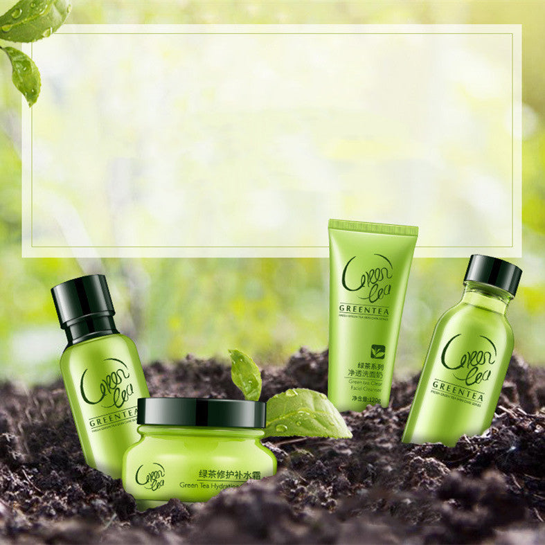 Four Green Tea Care products from the brand 'Green Tea' are arranged on dark soil with green leaves against a soft, blurred green background. The products include a bottle, a jar, and a tube, all in a vibrant green color with black lids and white branding.