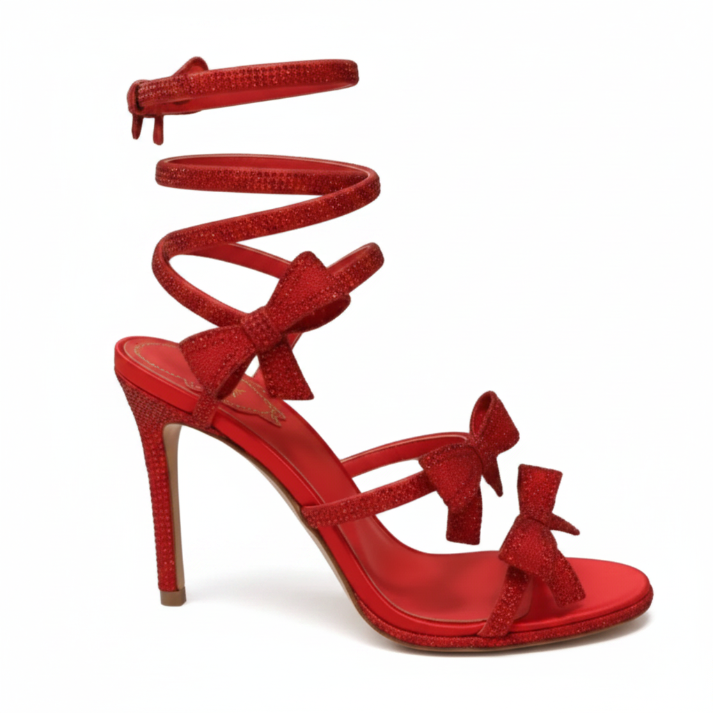 A red glittery high-heeled sandal with multiple bow details and an ankle strap, held by a hand wearing a black glove in front of a ribbed metallic background.