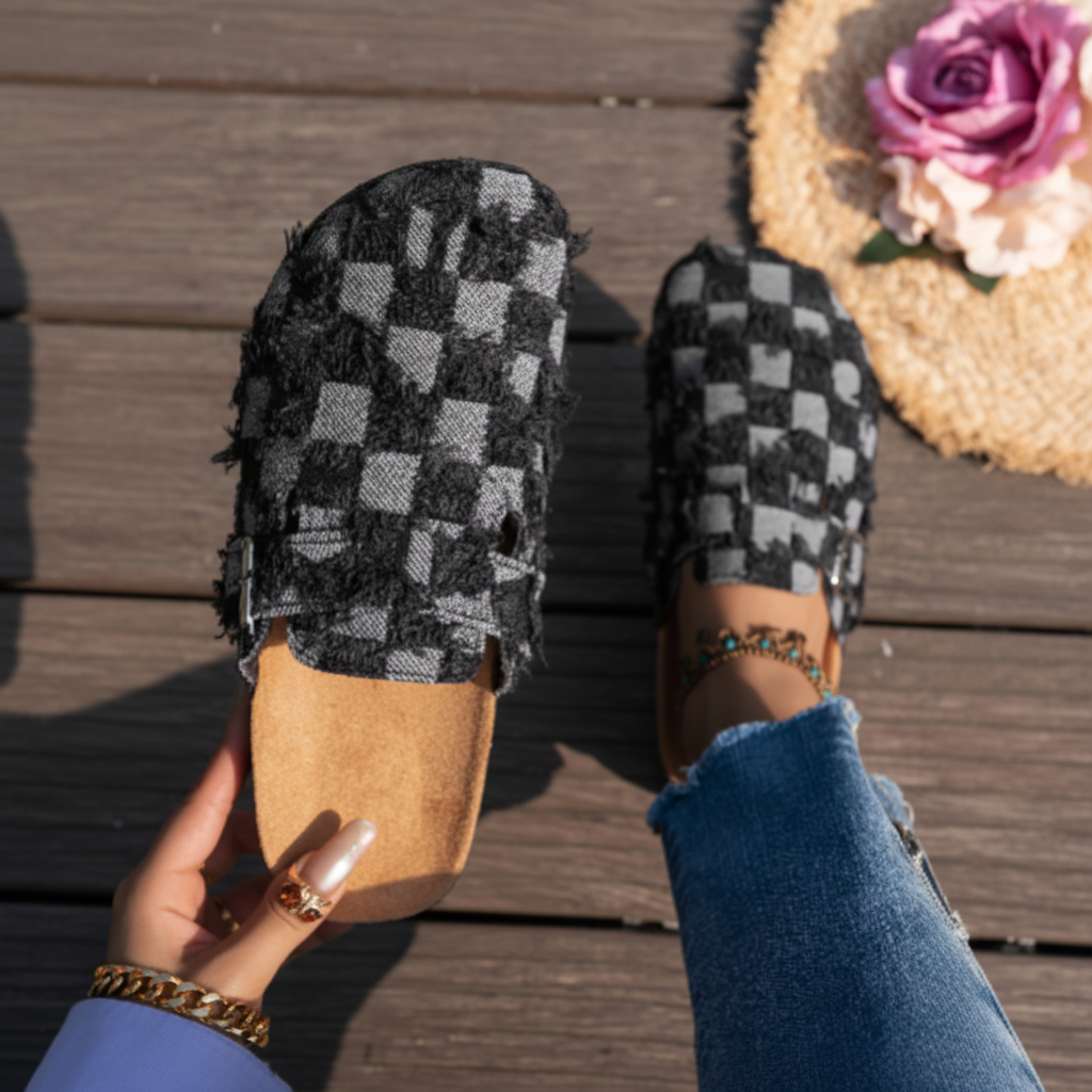 A person wearing a black and gray checkerboard patterned mule slipper with a tan suede-like footbed, shown holding the other slipper over a wooden deck. The person is wearing blue jeans and a gold anklet.
