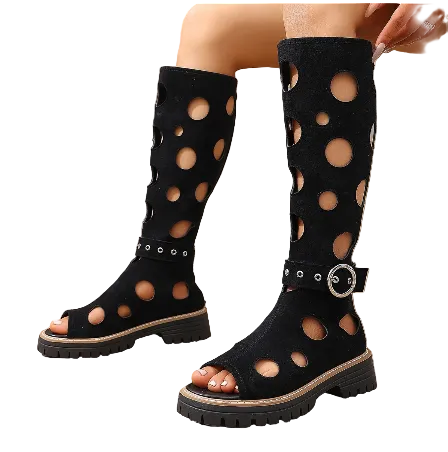 A pair of black knee-high cutout boots with a chunky lug sole and a decorative silver buckle strap, shown on a model's feet against a stone wall background.