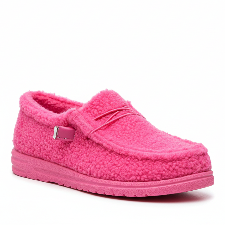 A person wearing a pair of bright pink fuzzy slip-on shoes, with one shoe being held up by a hand. The shoes have a textured fleece-like upper and a small decorative bow on the side.