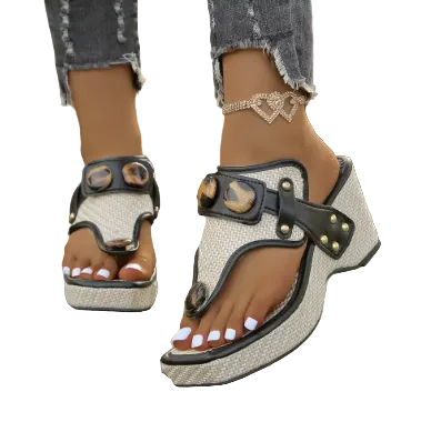 A pair of women's platform thong sandals with a textured beige woven upper, black faux-leather straps, and brown shell embellishments, shown on a model's feet standing on a wooden deck outdoors.