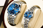 Two OLEVS wristwatches displayed on cream-colored cushion inserts. One watch is larger and features a blue dial with gold-tone details, while the other is smaller with a matching blue dial.