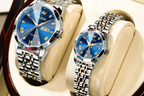 Two OLEVS wristwatches displayed on cream-colored watch cushions. One larger watch and one smaller watch, both with blue faces and silver-and-gold metal link bands, set against a dark wood and cream background.