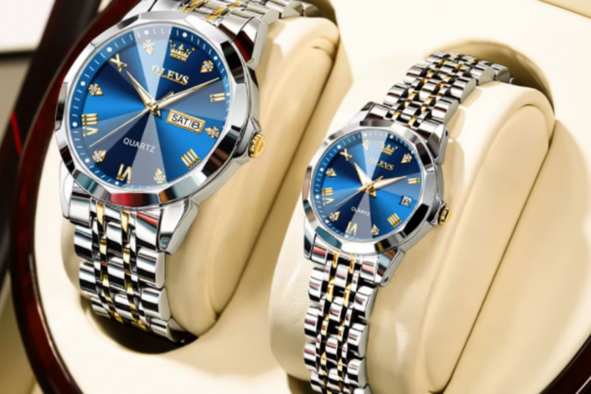 Two OLEVS wristwatches displayed on cream-colored watch cushions. One larger watch and one smaller watch, both with blue faces and silver-and-gold metal link bands, set against a dark wood and cream background.