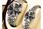 Two OLEVS men's analog watches displayed on cream-colored watch stands. The watches feature polished silver-tone stainless steel cases and multi-link metal bracelets with gold-tone accents, black dials with gold Roman numerals, and date windows.