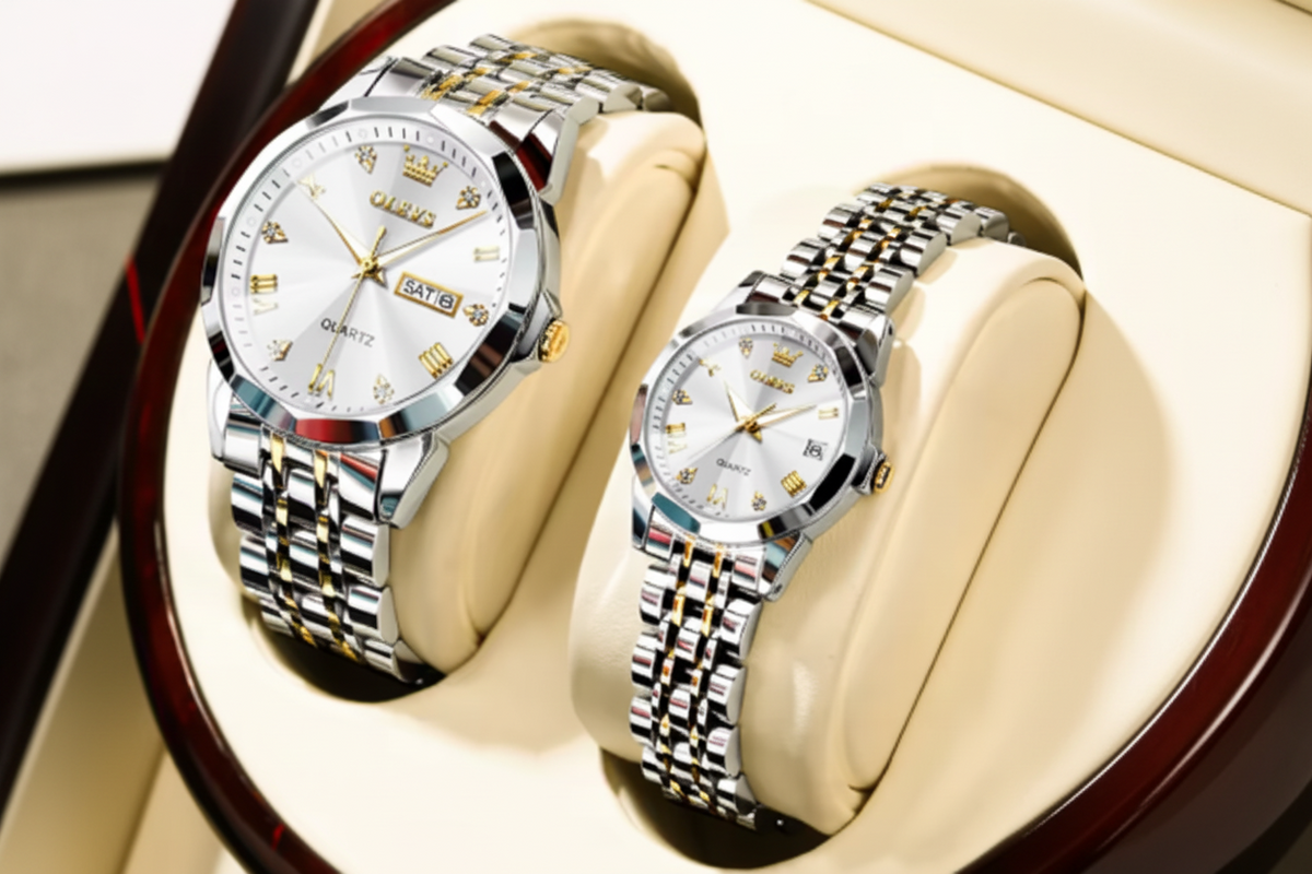 Two OLEVS men's quartz wristwatches with silver and gold-tone metal link bracelets and white dial faces, displayed on beige watch stands inside a watch box.