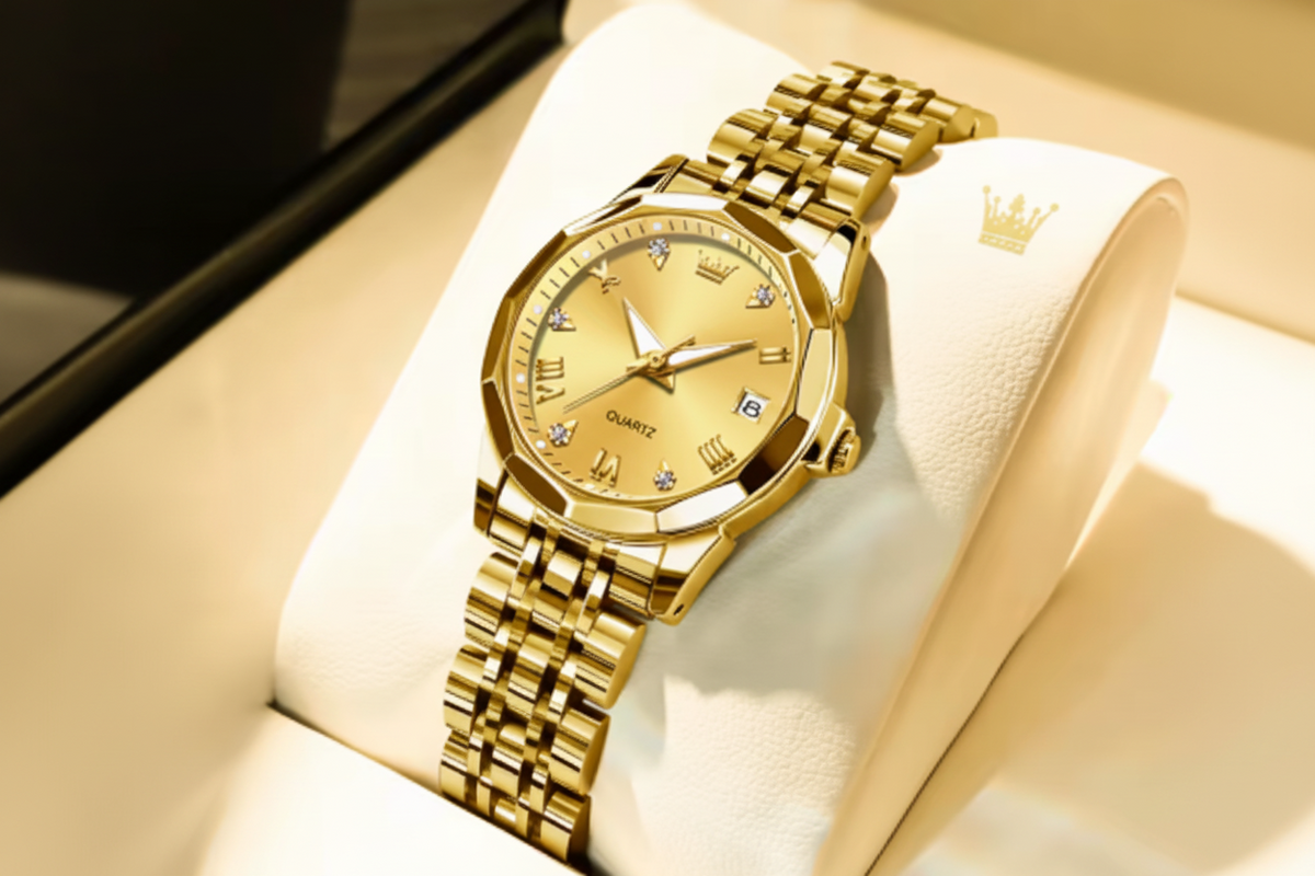 Gold-tone OLEVS women's quartz watch with a matching metal link bracelet, displayed on a cream-colored jewelry stand. The watch features a gold dial with crystal hour markers and a date window at 3 o'clock.