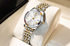 Olevs men's analog watch with a silver and gold-tone stainless steel link bracelet, a white dial with gold details, and a date window, displayed on a cream-colored watch cushion.
