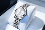A silver stainless steel wristwatch with a white dial and crystal glass, resting on a white display cushion. The brand name 'OLEVS' is visible in a red logo overlay in the top-left corner.