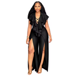 A woman modeling a black ruffled jumpsuit with a high leg slit and strappy lace-up neckline, paired with gold high-heeled sandals, isolated on a white background.
