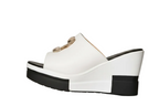 White platform wedge sandals with cutout straps and black soles, shown on a model's feet in a minimalist indoor setting.