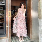 A woman wearing a light pink floral spaghetti-strap maxi dress with 3D floral appliqué and layered ruffle details, standing in a rustic wooden doorway.