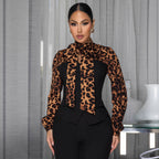 A woman models a leopard print long-sleeve blouse with a high neck and bow detail, paired with black pants, set against a modern interior background with sheer curtains and a white vase.