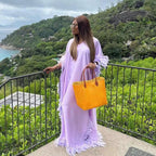 Woman in a lavender dress holding an orange bag with a scenic background