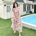 A woman stands outdoors in front of a cream-colored building with a swimming pool, wearing a sleeveless pink floral maxi dress with layered ruffle details. She is holding a small white handbag and a phone.