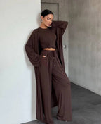 A woman models a matching brown two-piece lounge set consisting of a long-sleeve crop top and high-waisted wide-leg pants, posed in a minimalist room with concrete walls.