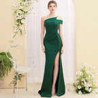 Woman wearing a green one-shoulder dress with a slit, standing in a decorated room with flowers and a chair.